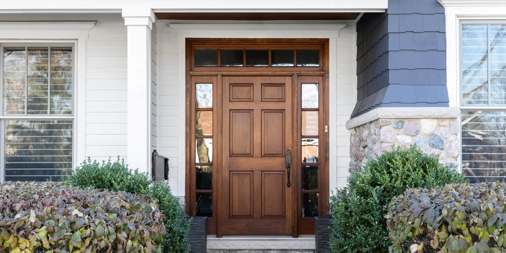 A wooden front door, surrounded by windows, with white, blue, and stone siding.